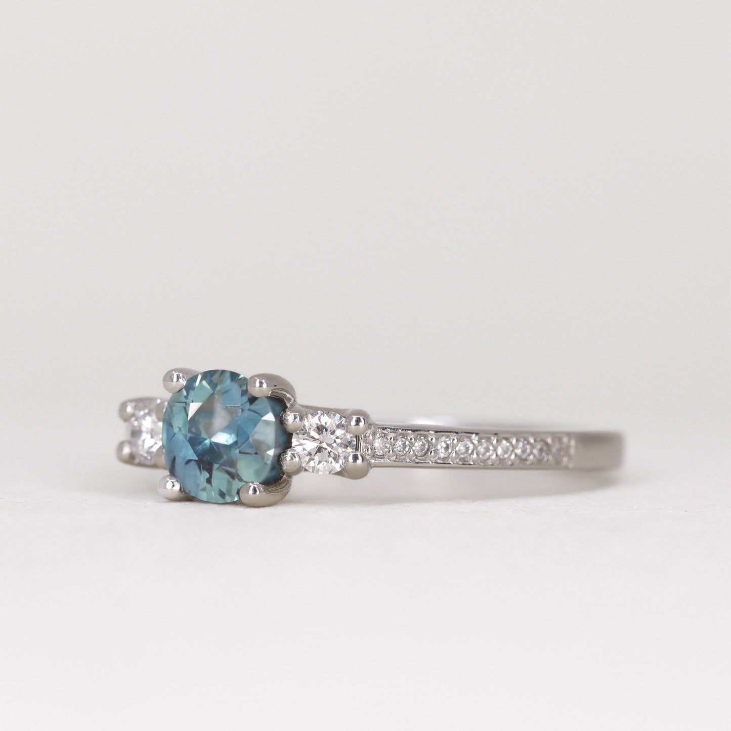 Round cut teal sapphire trilogy engagement ring with pave band